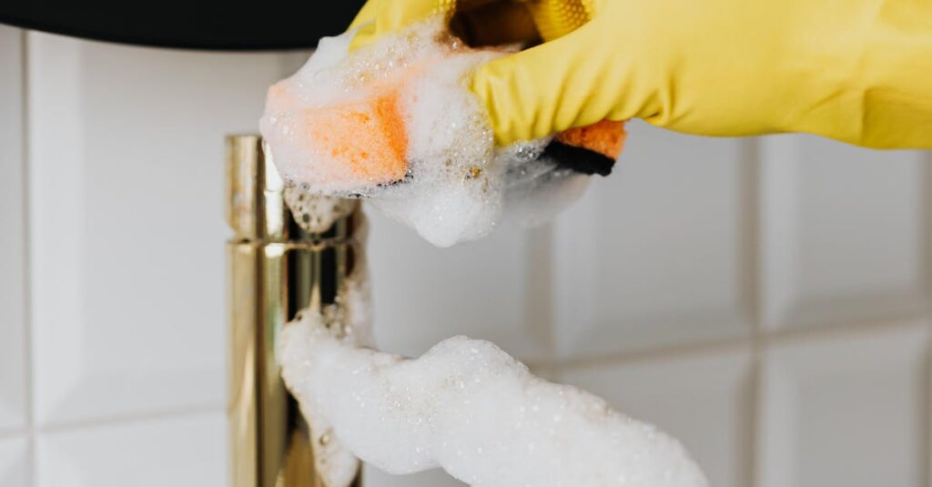 Close-up of a hand in a yellow glove cleaning a bathroom faucet with foam.